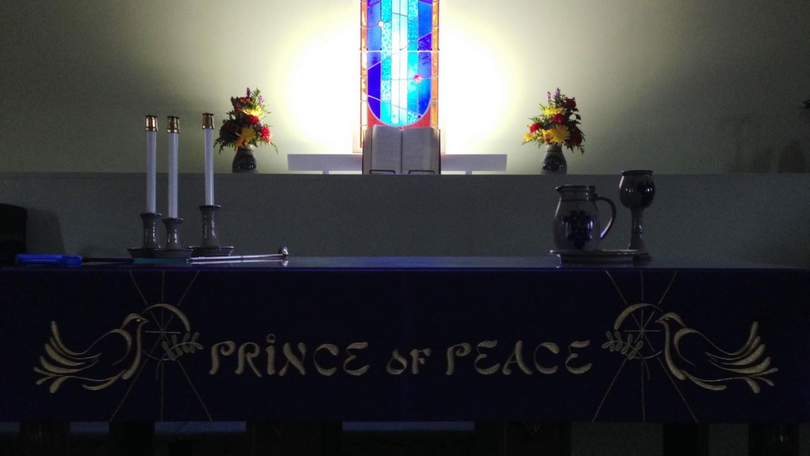 Church altar with “Prince of Peace” banner, candles, flowers, and stained glass centerpiece