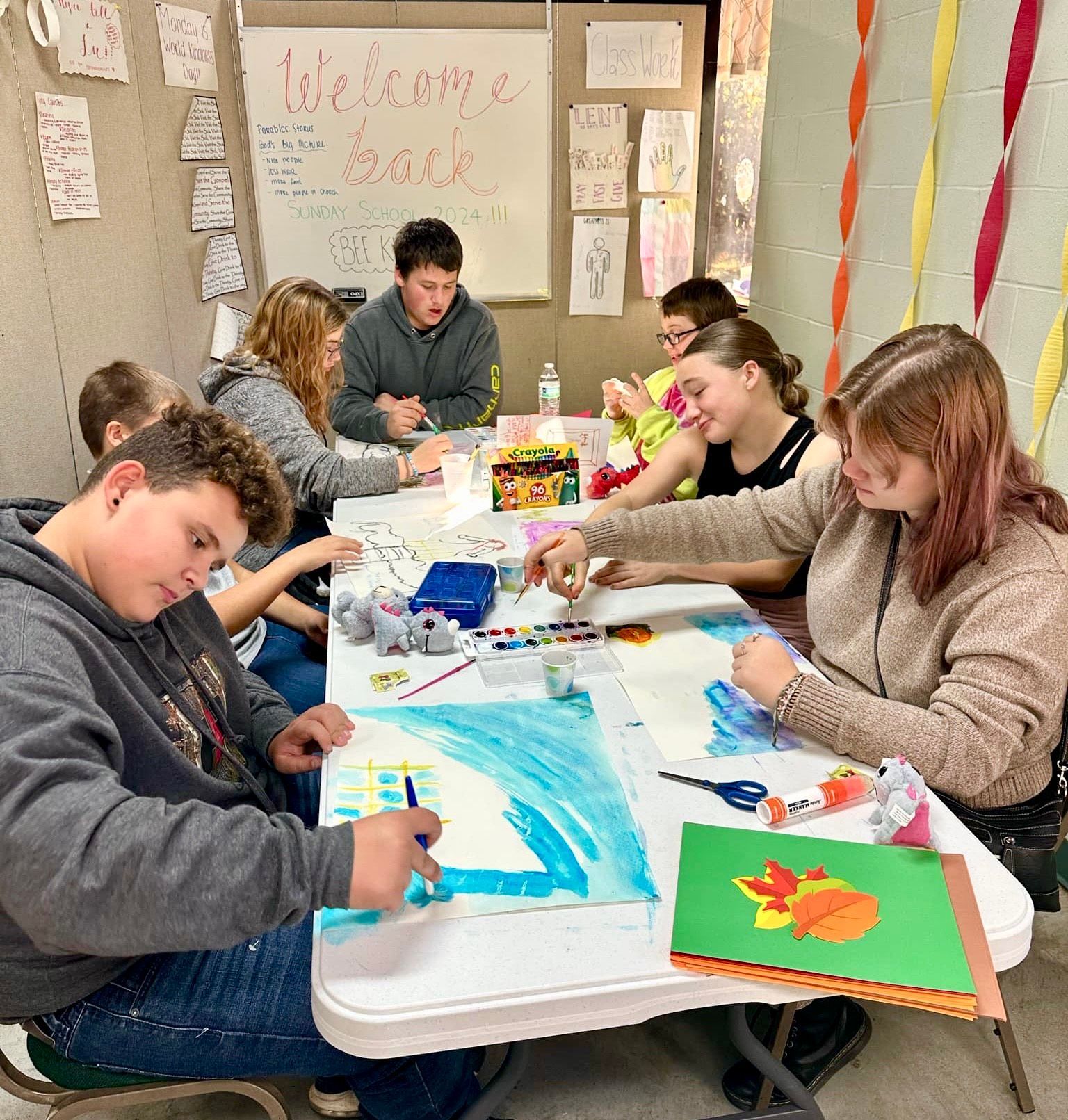 People painting at a classroom table with colorful art supplies and paper decorations on the walls