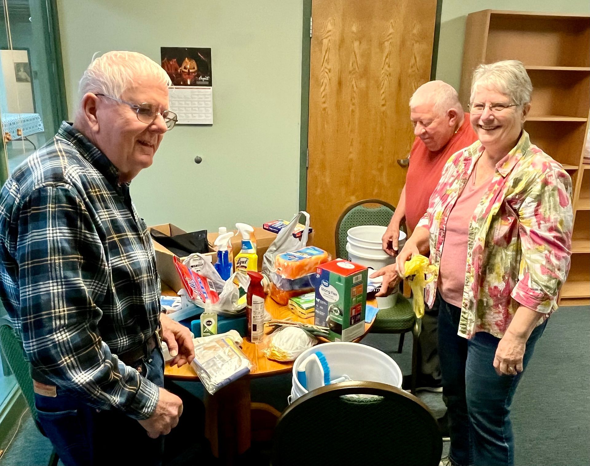 Three seniors stand by a snack table in a room, smiling beside drinks and food items.