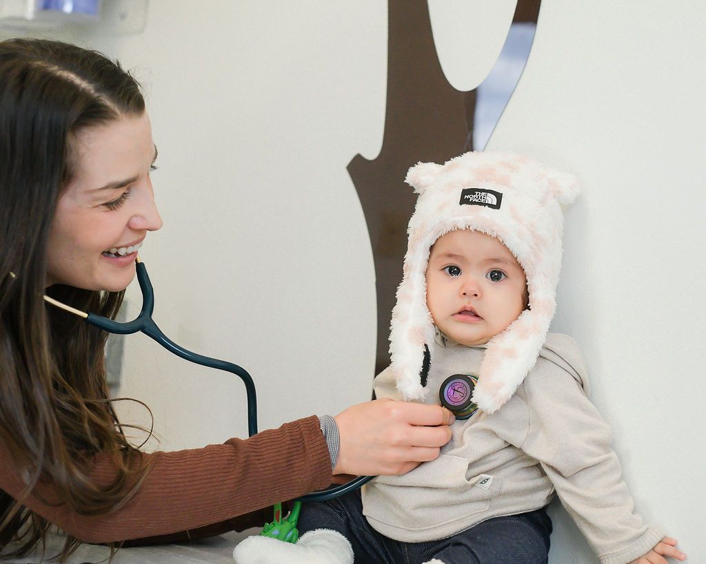 A pediatrician is listening to a baby's heart using a stethoscope, the baby is wearing a fuzzy hat