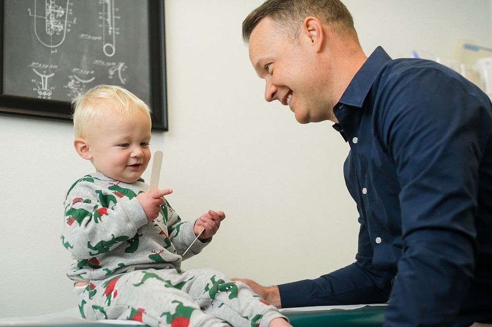 A smiling doctor sitting with a toddler in a patterned outfit holding a wooden tongue depressor in a clinical office.