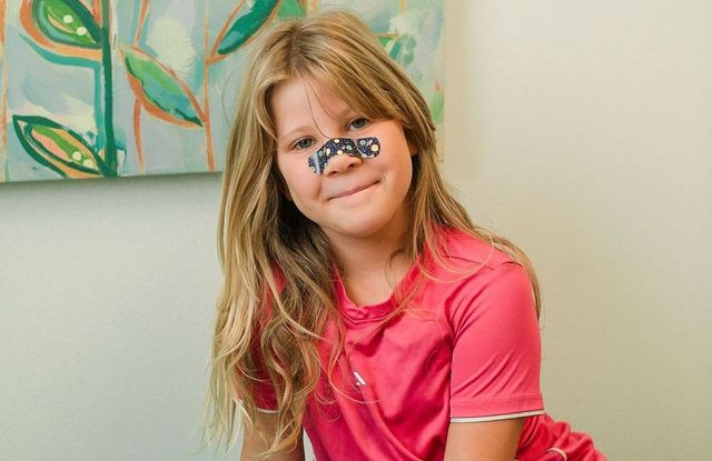 Child in a pink shirt smiling, with a small toy car on her nose, indoors near a colorful painting