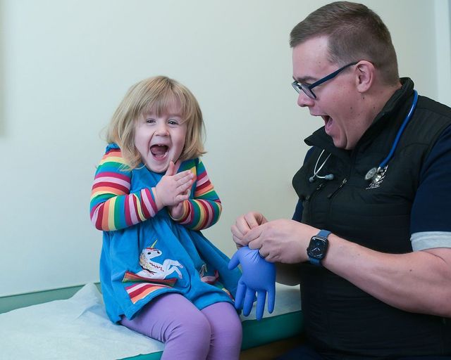 Child sitting on exam table receiving a blue glove from a smiling clinician in a medical office