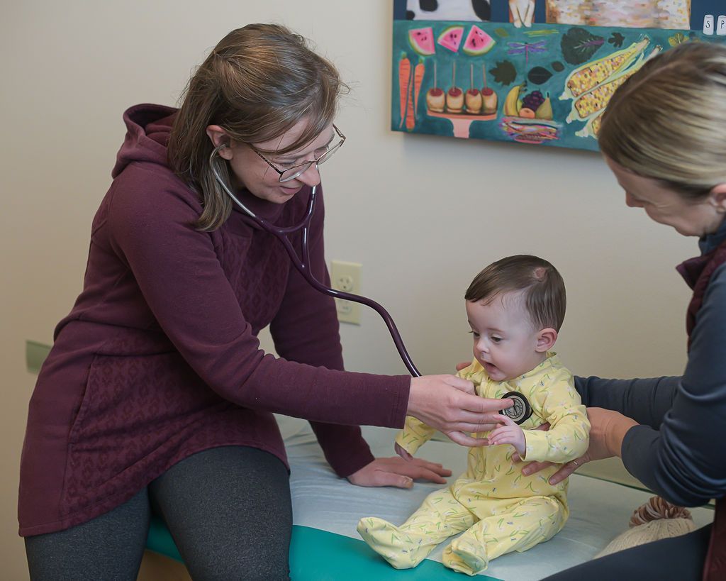 Doctor examines baby's chest with stethoscope in doctor's office. Another person assists.