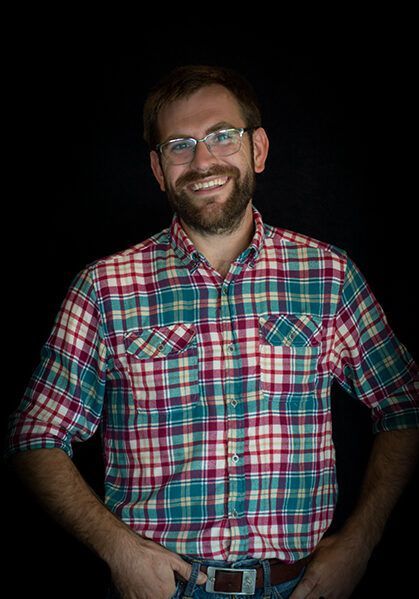 Man with a beard and glasses wearing a plaid shirt, smiling, hands in pockets, black background.