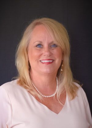 Blonde woman with a pearl necklace smiles at the camera; she is wearing a light pink blouse.