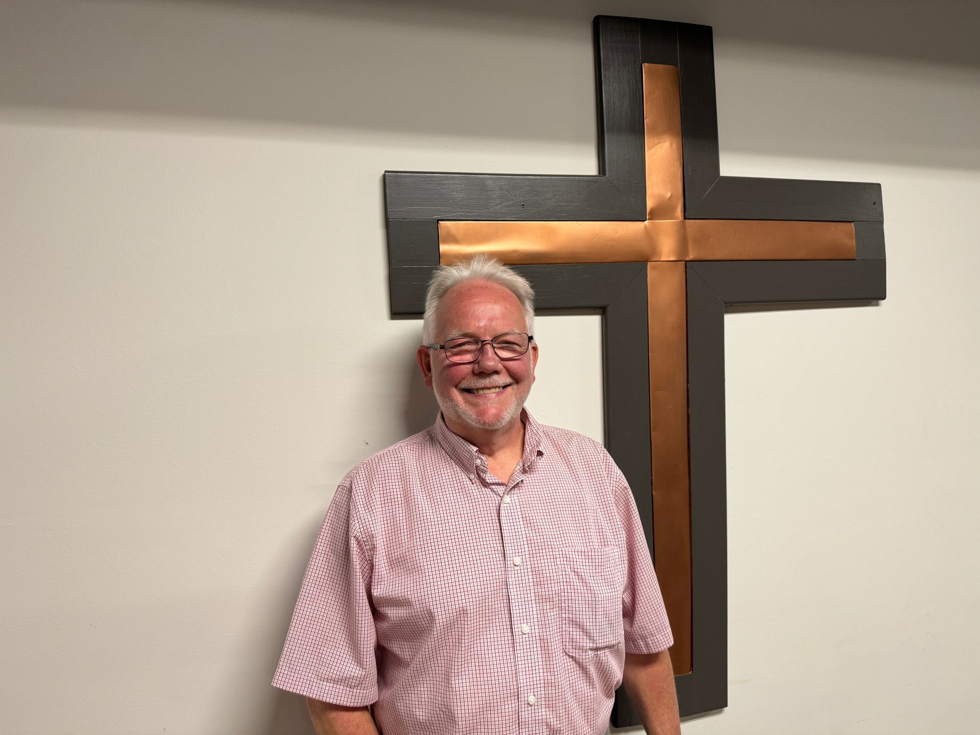Man with glasses smiles, stands in front of a copper and dark gray cross on a white wall.