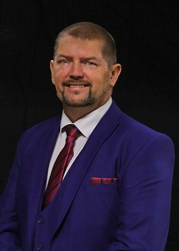 Man in a suit, smiling, with a goatee, set against a black background.
