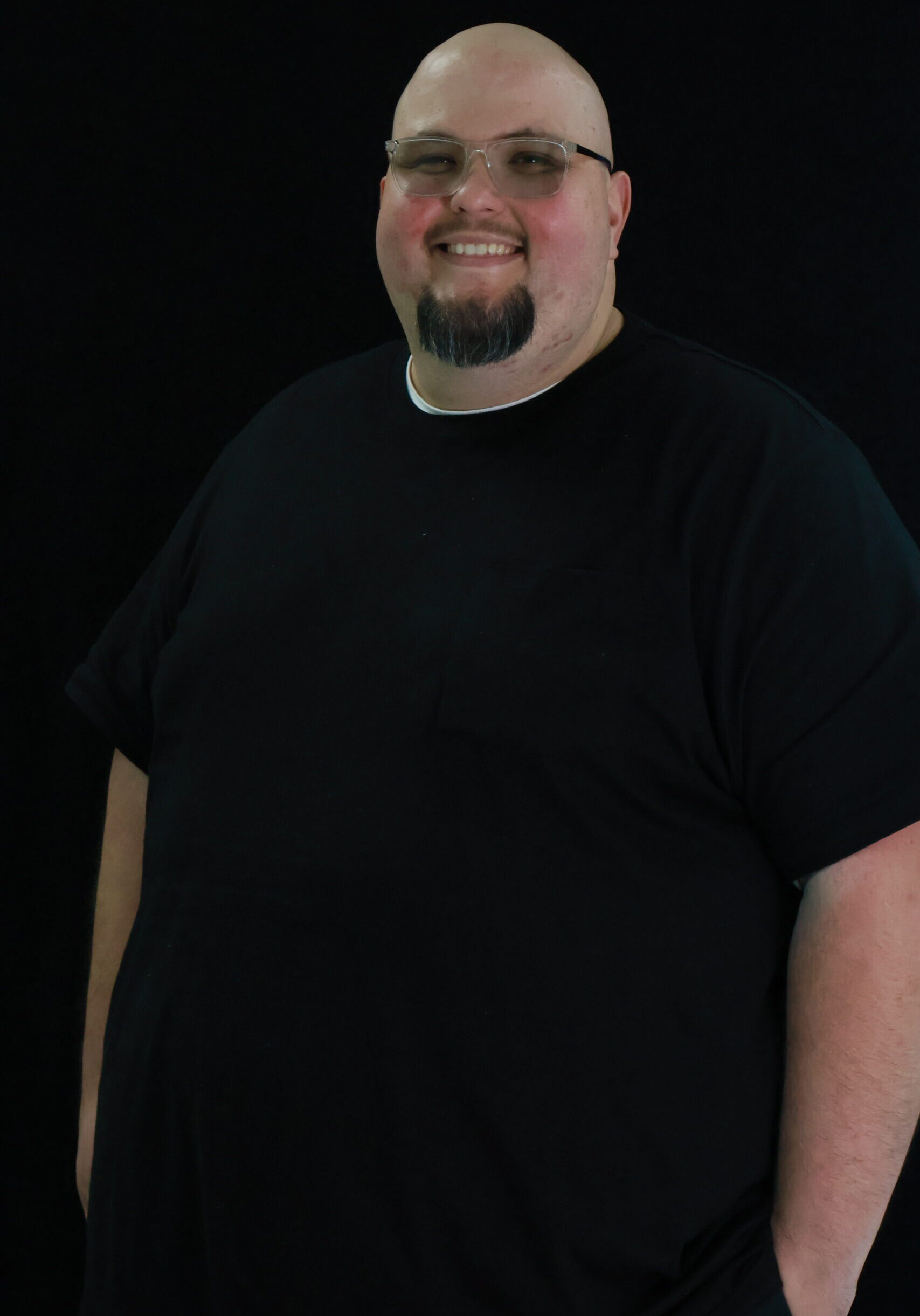 Man in black t-shirt and glasses, smiling with a trimmed beard, standing against a black backdrop.