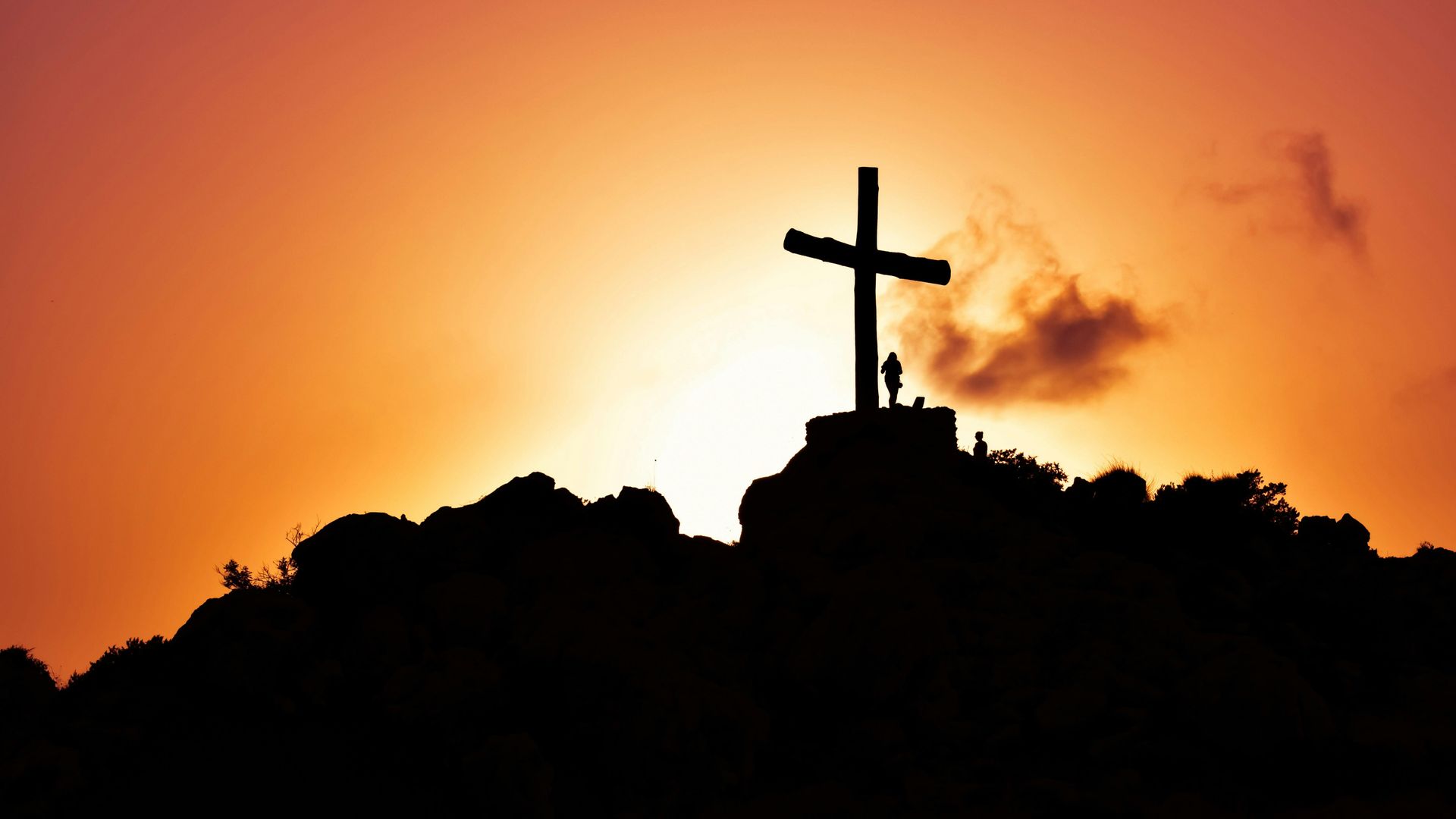 Silhouette of a cross on a hilltop against an orange sunset sky.