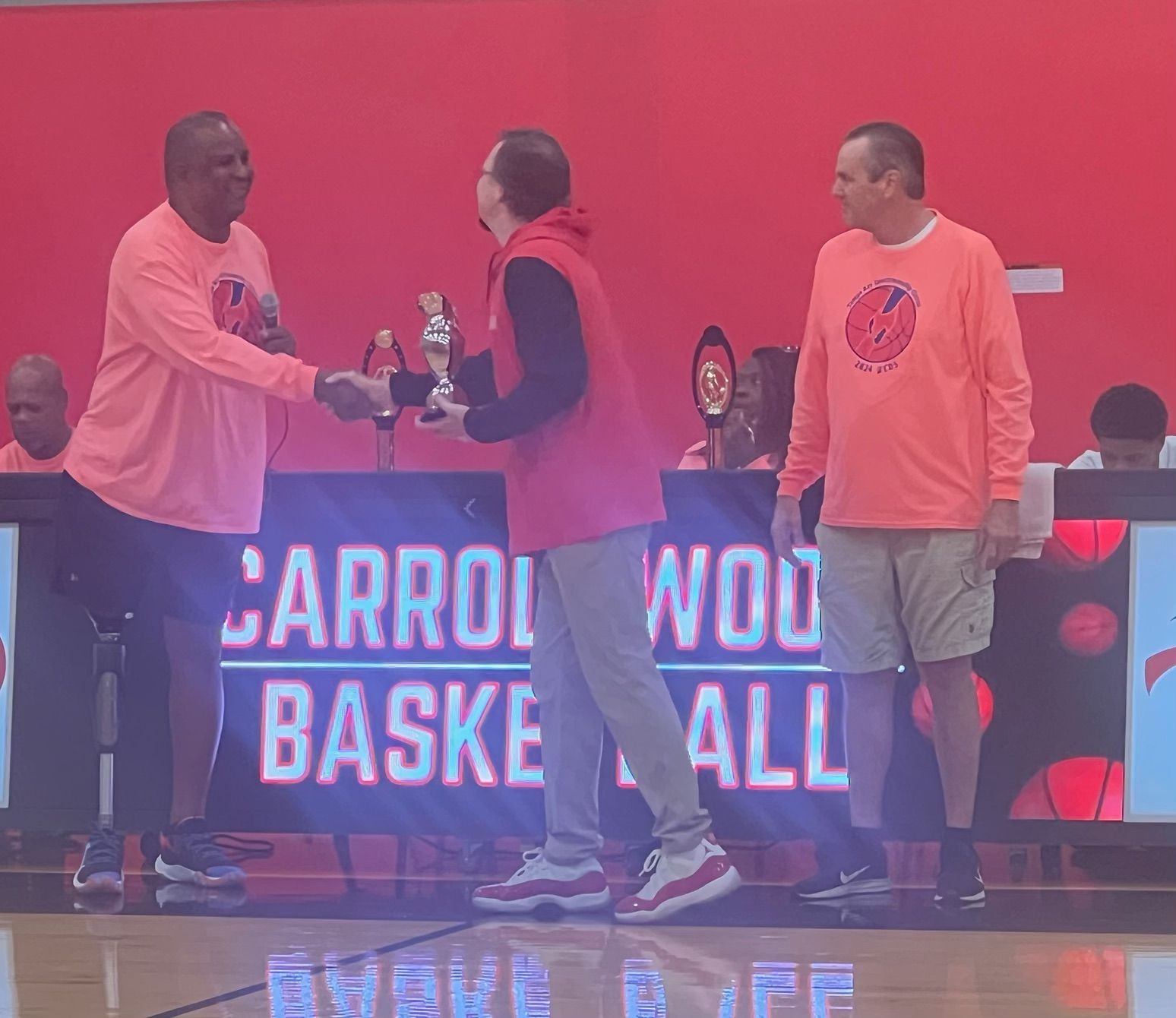 Two men shake hands as a third presents a trophy on a basketball court.