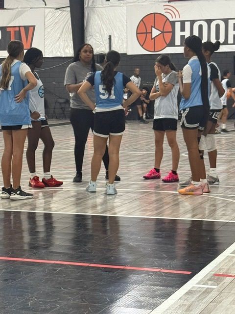 Basketball team huddles on court, coach talks; players wear blue and white jerseys, black shorts.