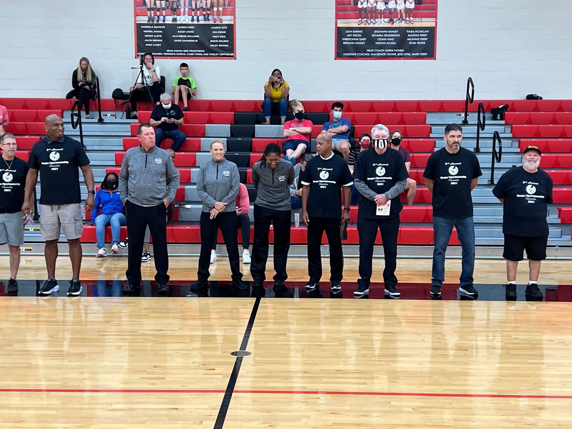 Coaches stand on a basketball court in front of bleachers with spectators. They wear matching black shirts.