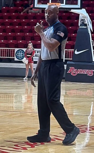 Basketball referee giving a thumbs up on court. Wearing black pants, striped shirt, and holding a whistle.