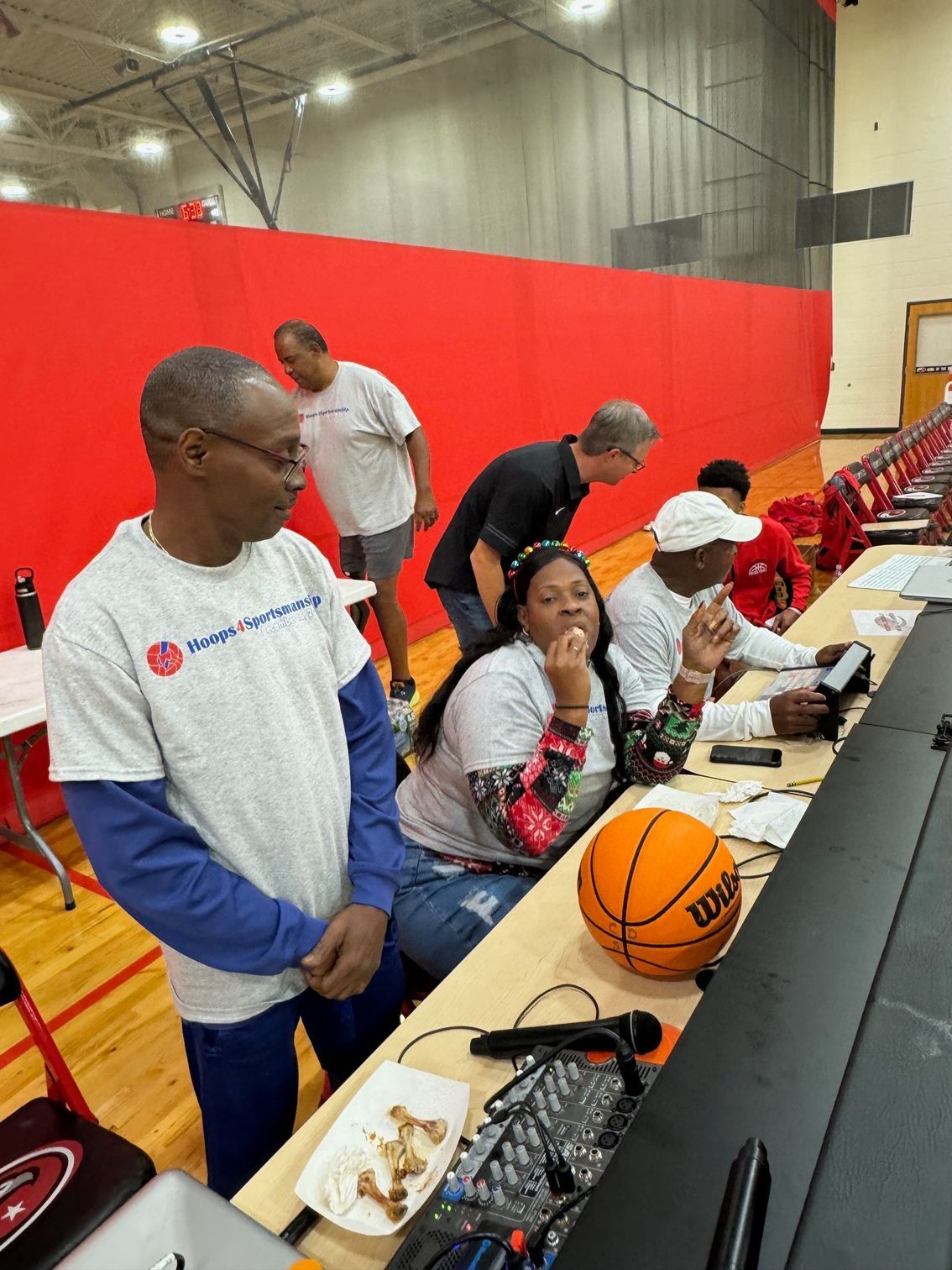 Basketball court with people at a scorer's table; one man watches intently. A woman gestures.