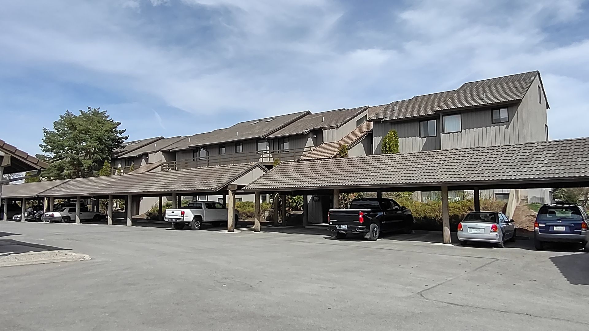 A row of cars are parked under a covered parking lot in front of a building.