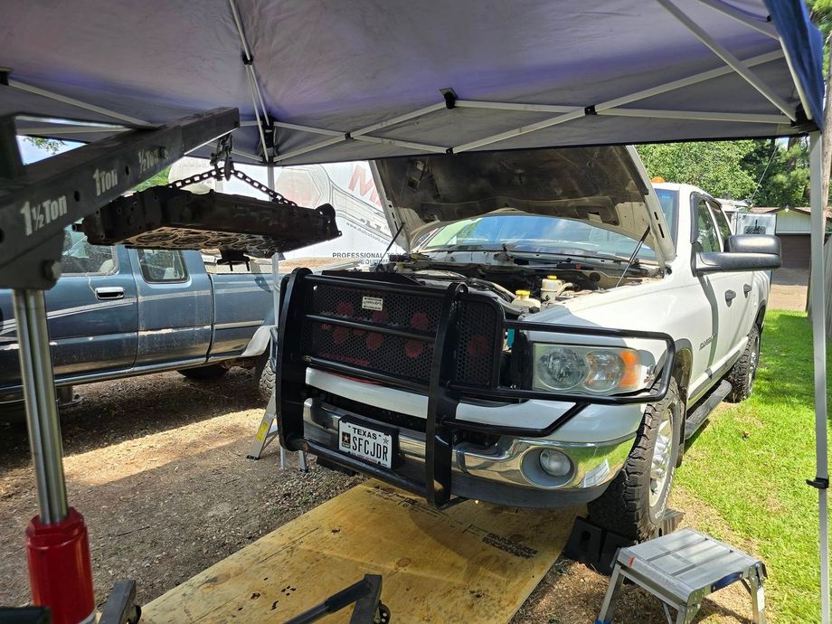 White pickup truck with front-end damage under a canopy, parked on a lift in a repair area