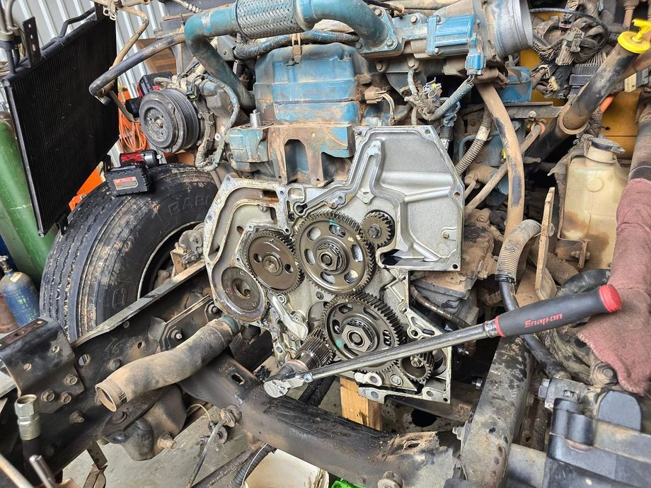 Car engine with front cover removed, exposing gears and belts; mechanic’s hand holds a wrench.