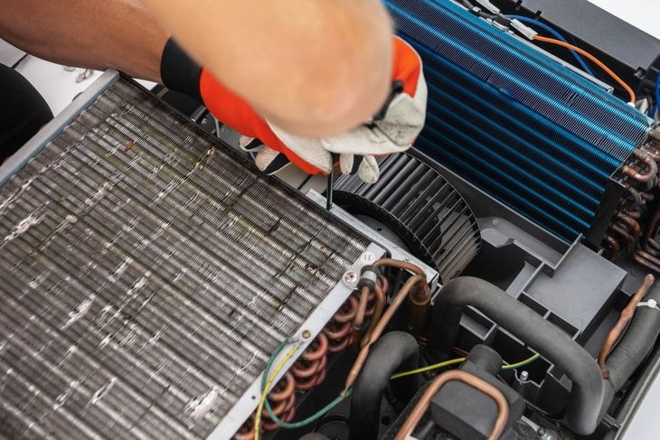 Hands in red gloves cleaning an air conditioner unit with exposed coils and wiring inside.