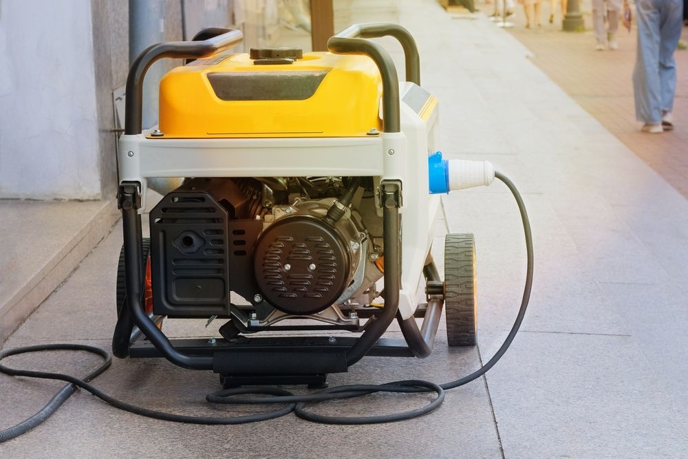 Portable generator on a sidewalk with a yellow fuel tank and coiled cords nearby.