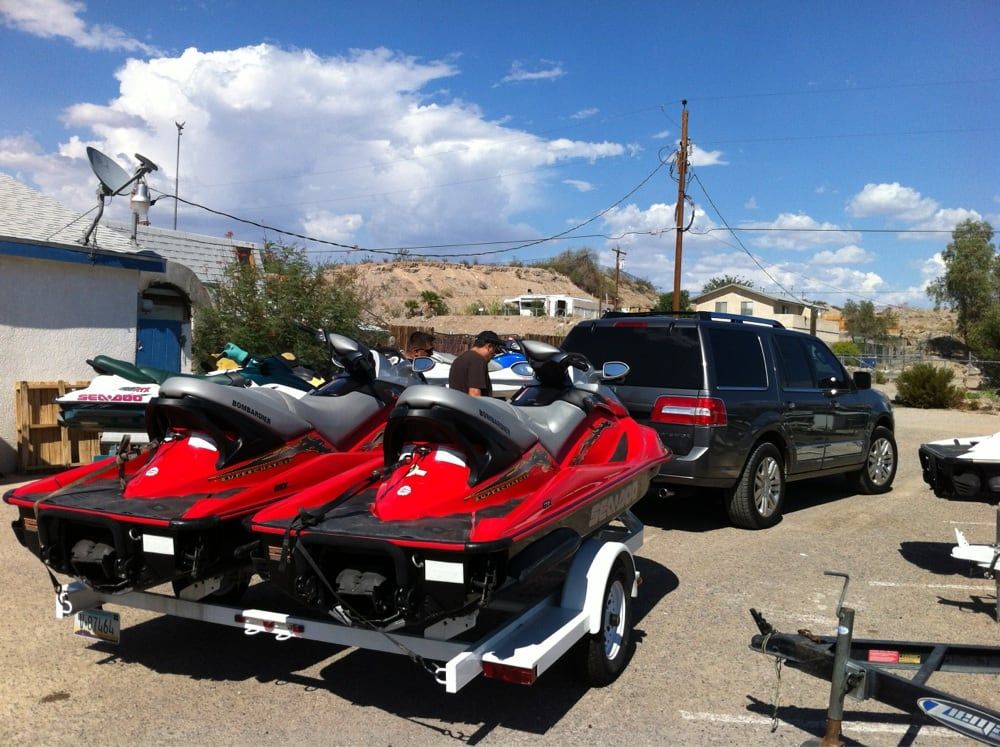 Rent from Dave's Wet & Wild Two red jet skis on a trailer beside a black SUV in a dusty lot under a blue sky