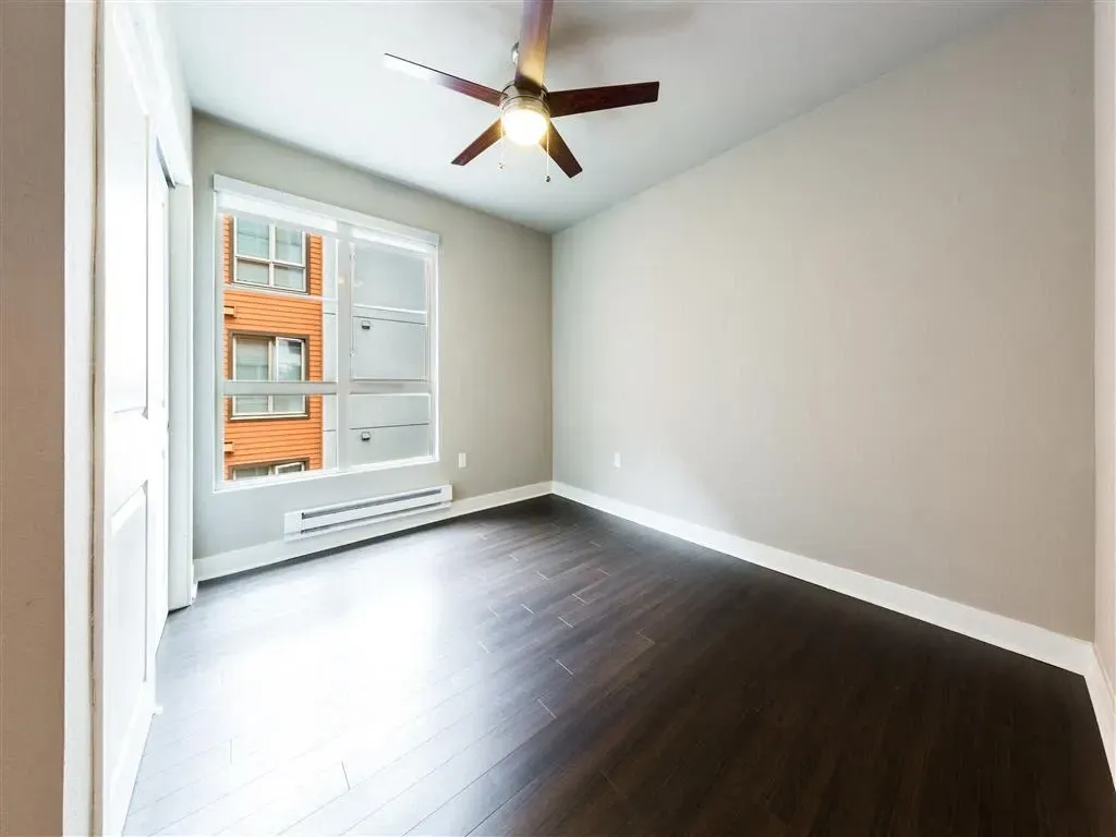 Empty bedroom with dark wood floors, a ceiling fan, and a window showing an adjacent building.