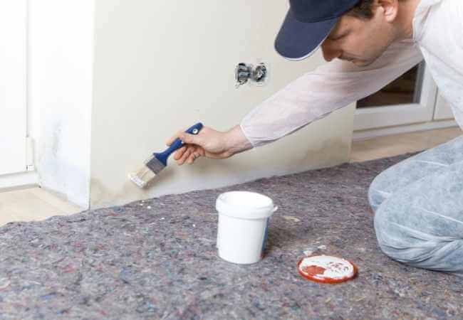 Una persona pinta la esquina de una habitación con una brocha; hay un cubo de pintura blanca en el suelo, que cubre el piso inferior.
