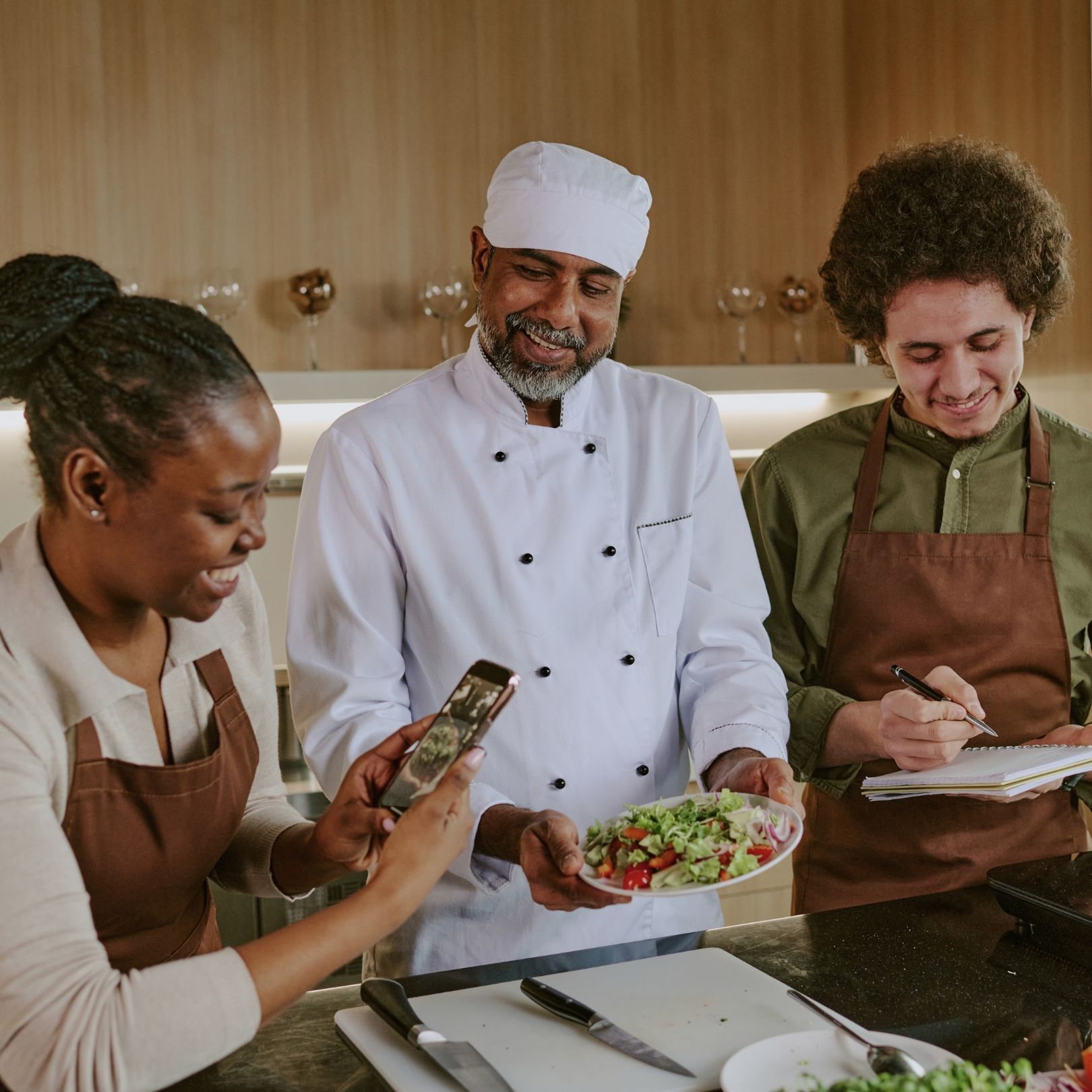 Three people cooking together in a kitchen, one holding a phone while another plates a salad.