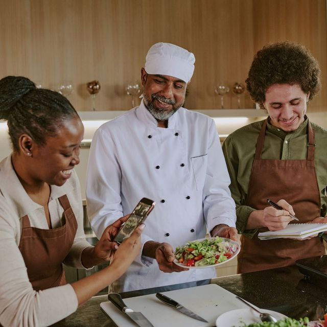 Three people cooking together in a kitchen, one holding a phone while another plates a salad.