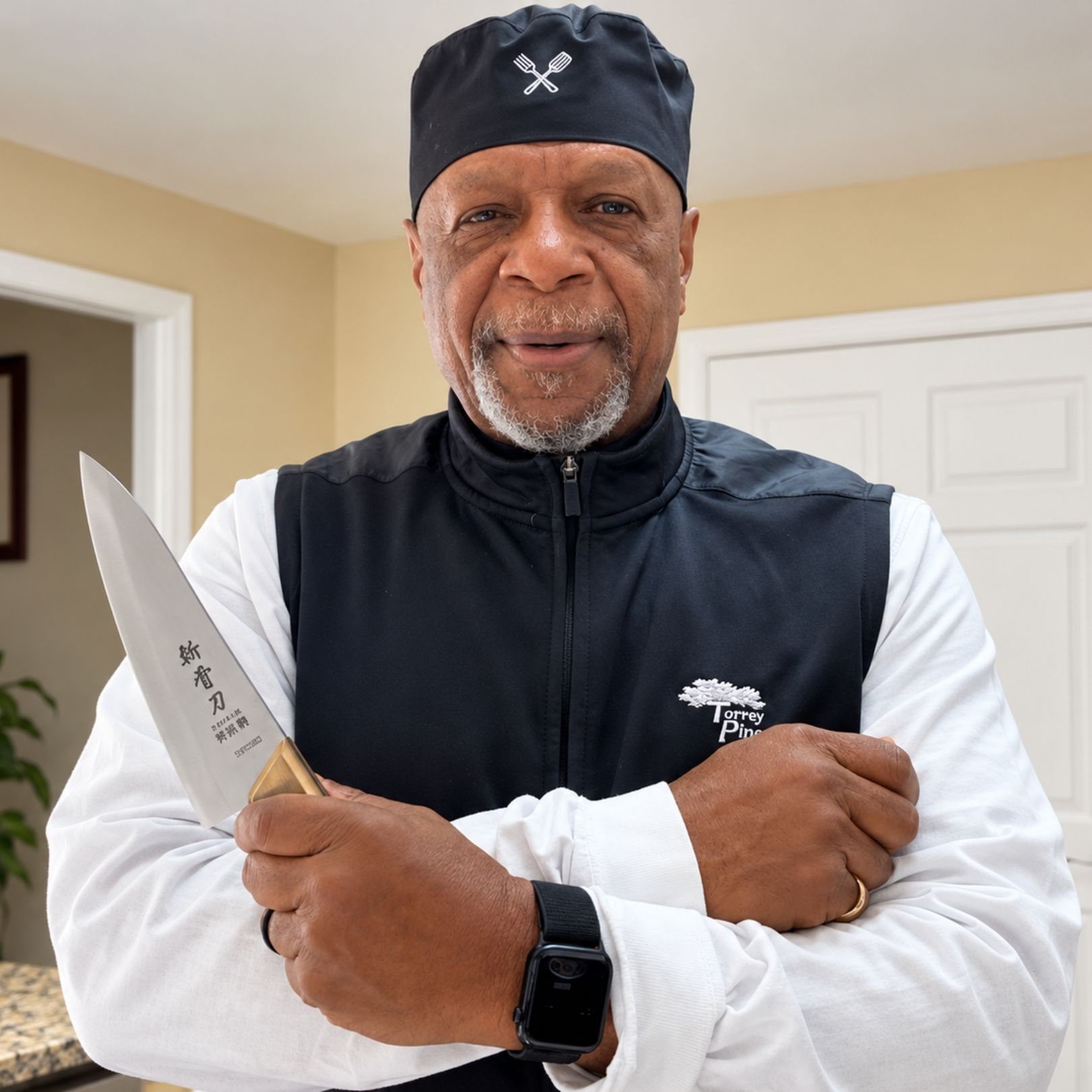Man standing indoors holding a chef’s knife, wearing a black cap, vest, and white shirt.