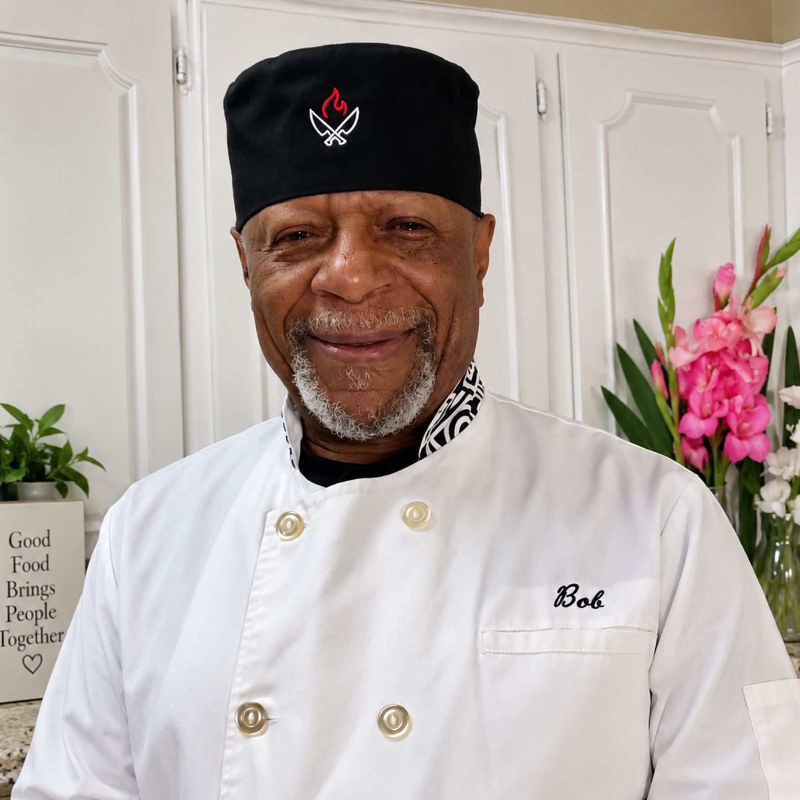 Smiling chef in white uniform and black hat standing in a kitchen with pink flowers in the background