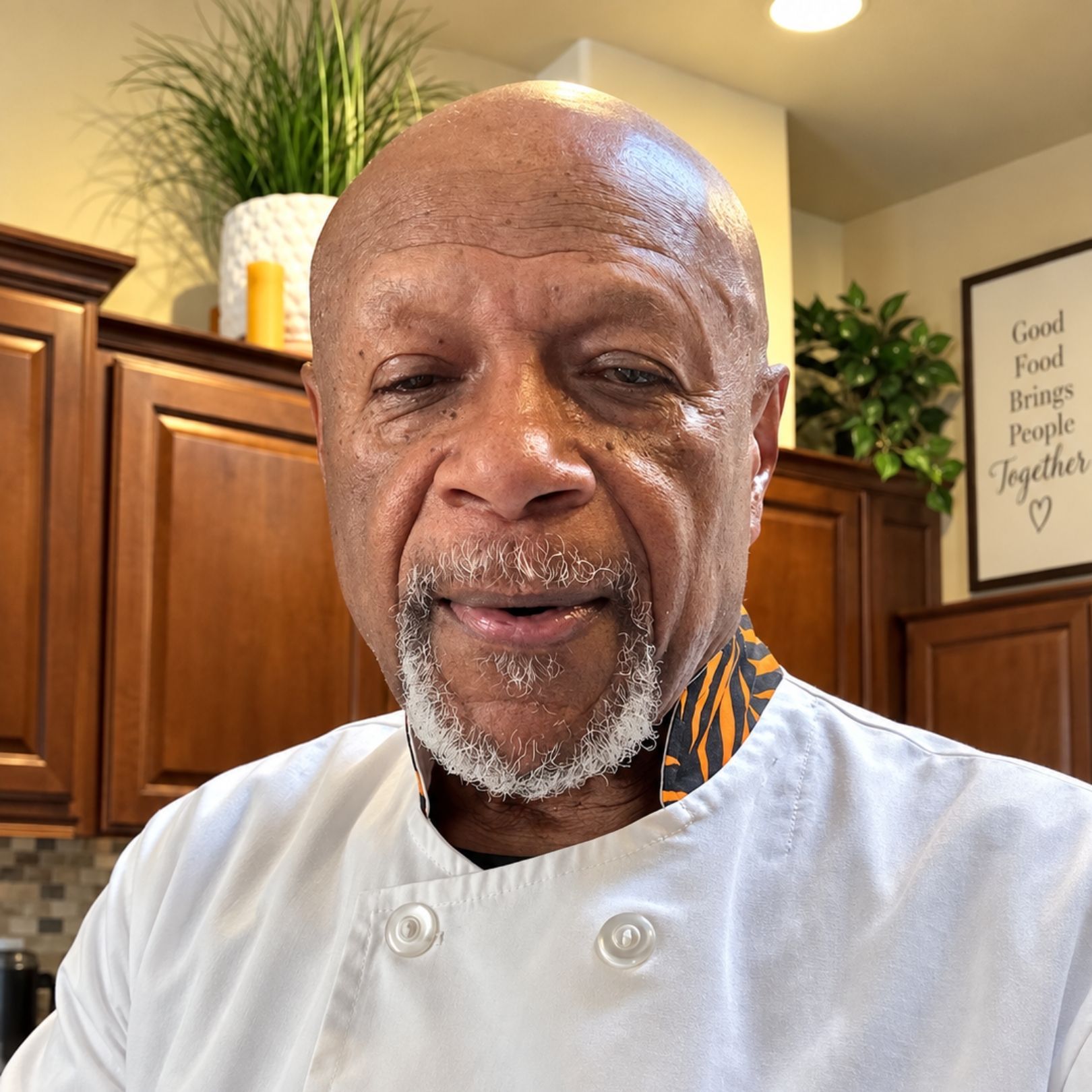 Smiling chef in a white jacket standing in a warm kitchen with wooden cabinets and a wall sign.