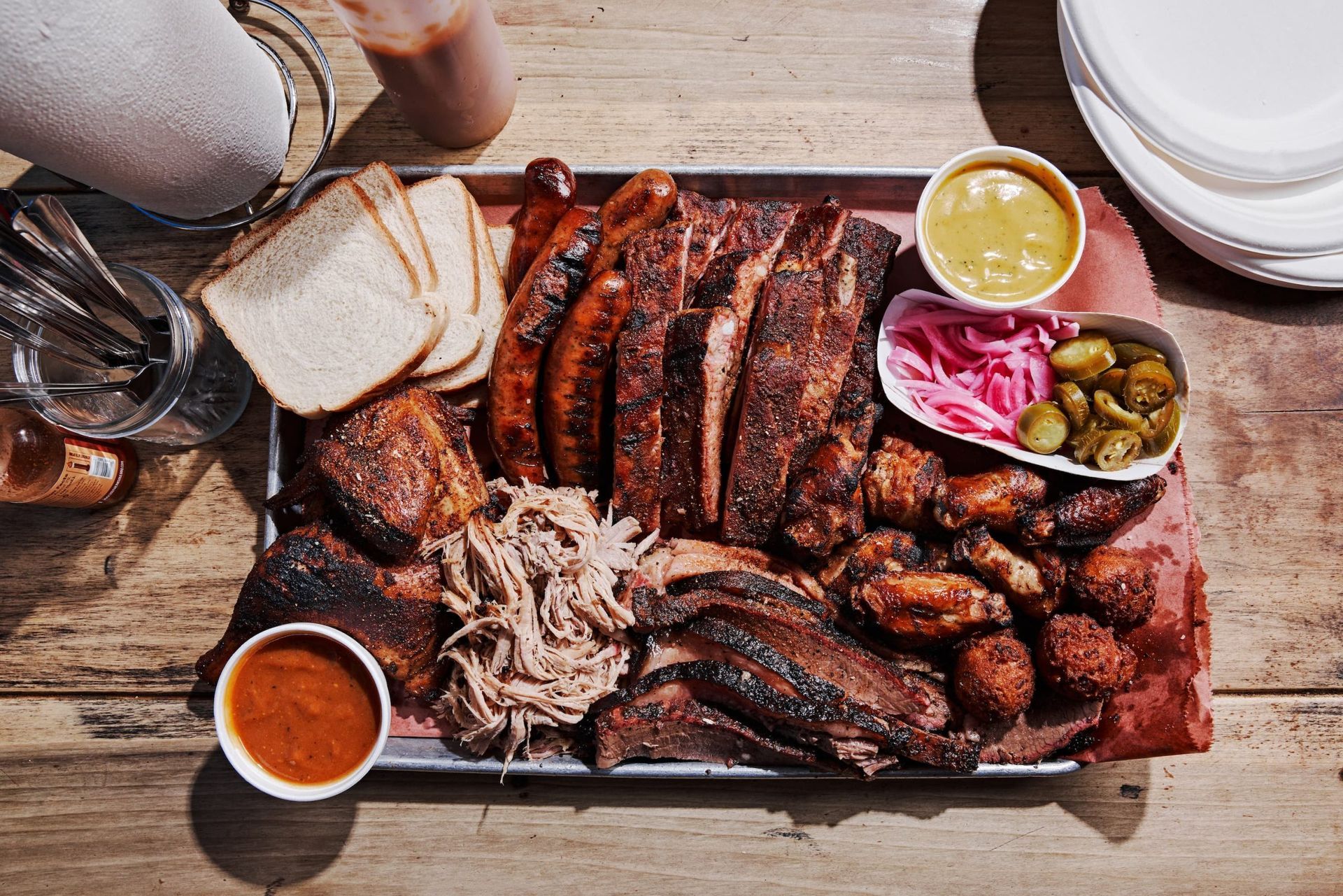 Tray of barbecue meats with bread, pickles, onions, and dipping sauces on a wooden table