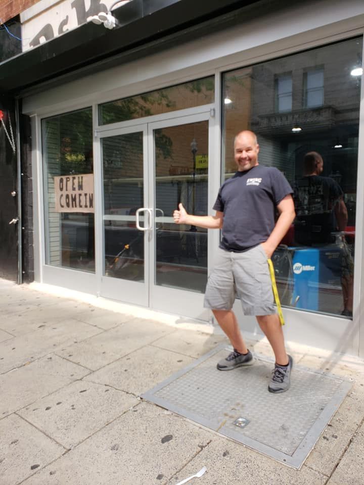 Man standing outside a storefront, holding the door open and smiling in sunlight.