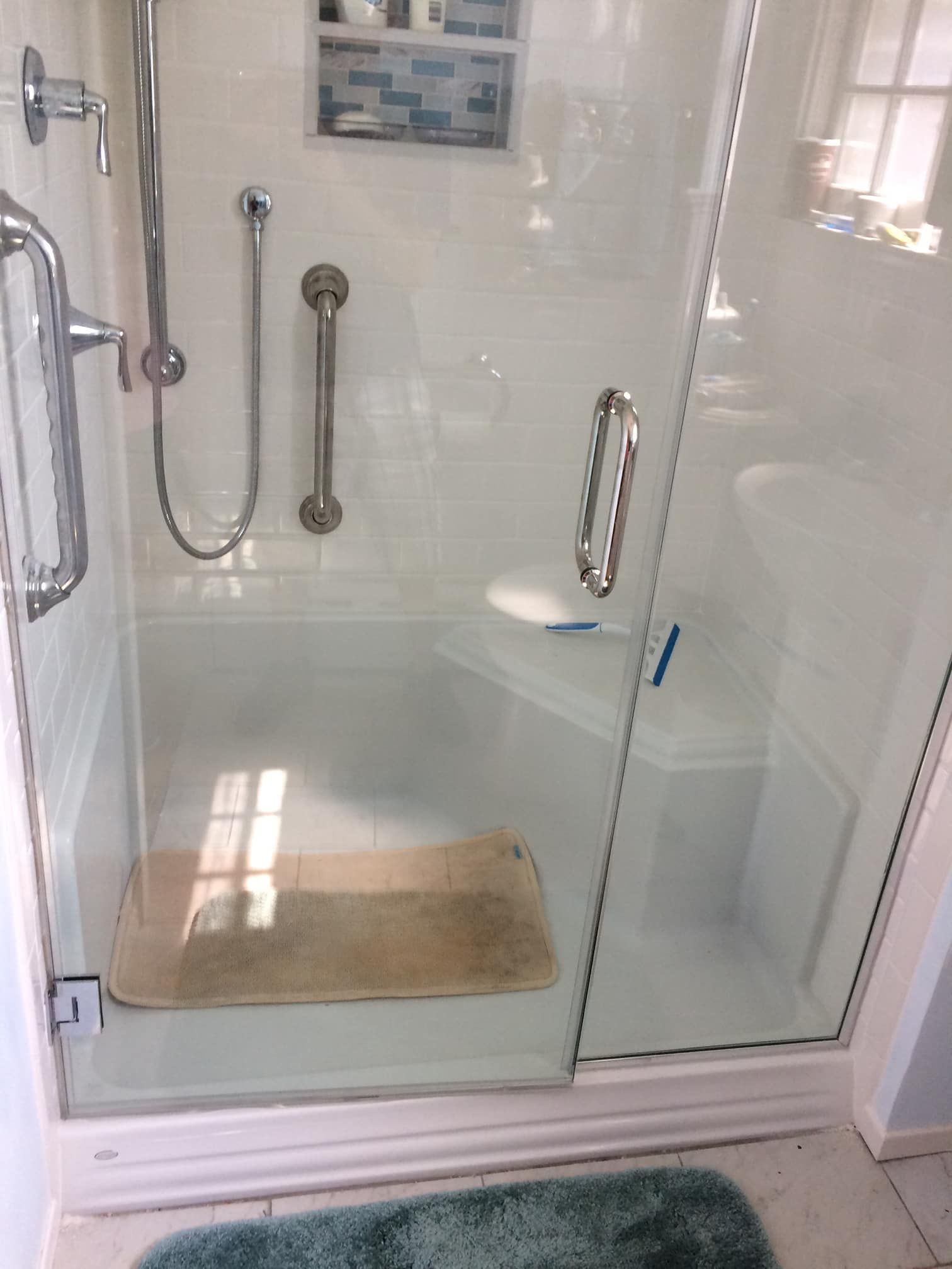Glass shower stall with chrome grab bars and a beige bath mat in a bright bathroom