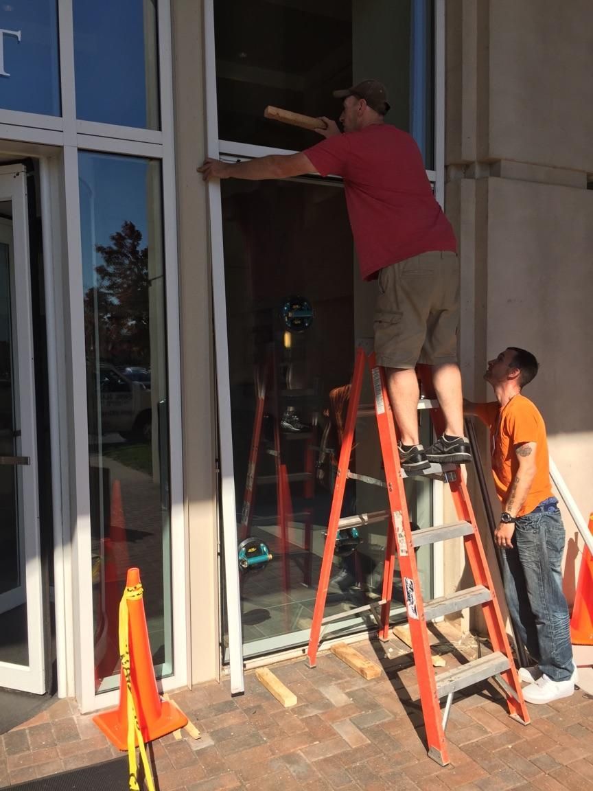 Two workers on ladders installing or cleaning a storefront window beside orange safety cones.