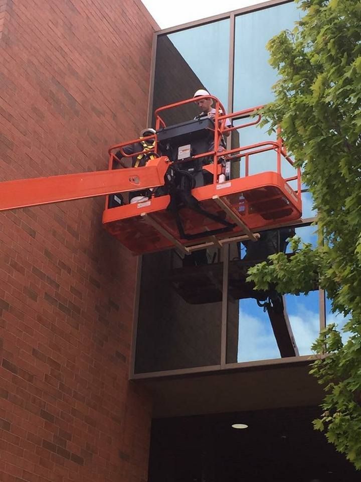 Worker in an orange lift repairing a brick building beside reflective windows.