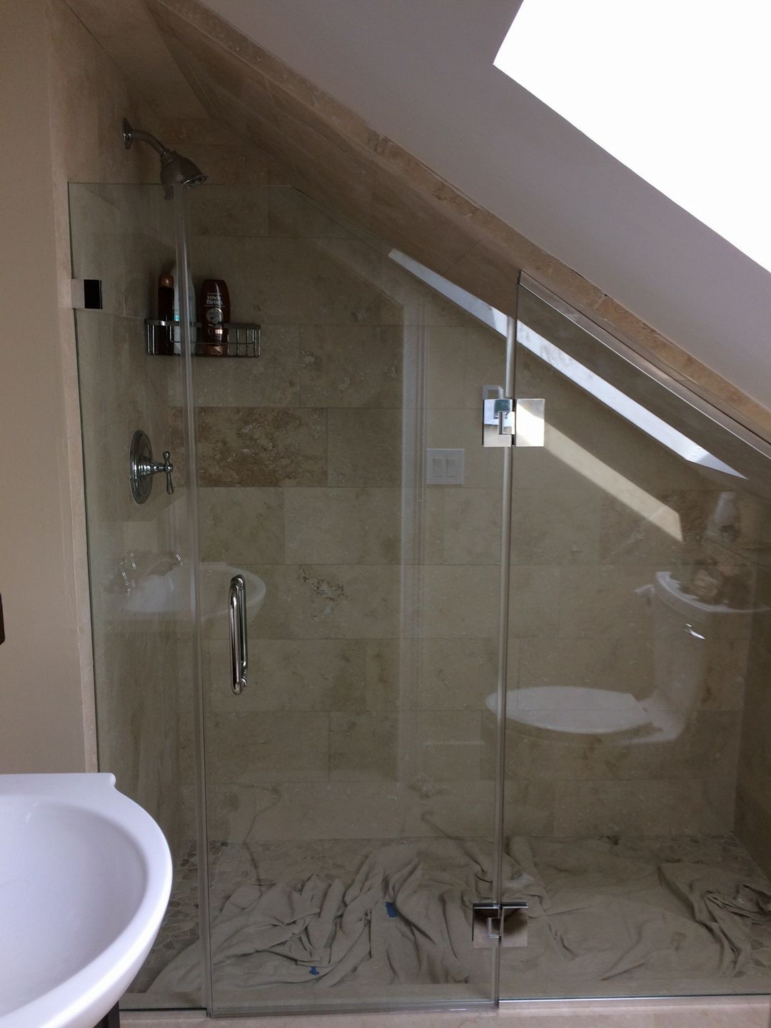 Glass shower under slanted ceiling with beige tile and a towel on the floor