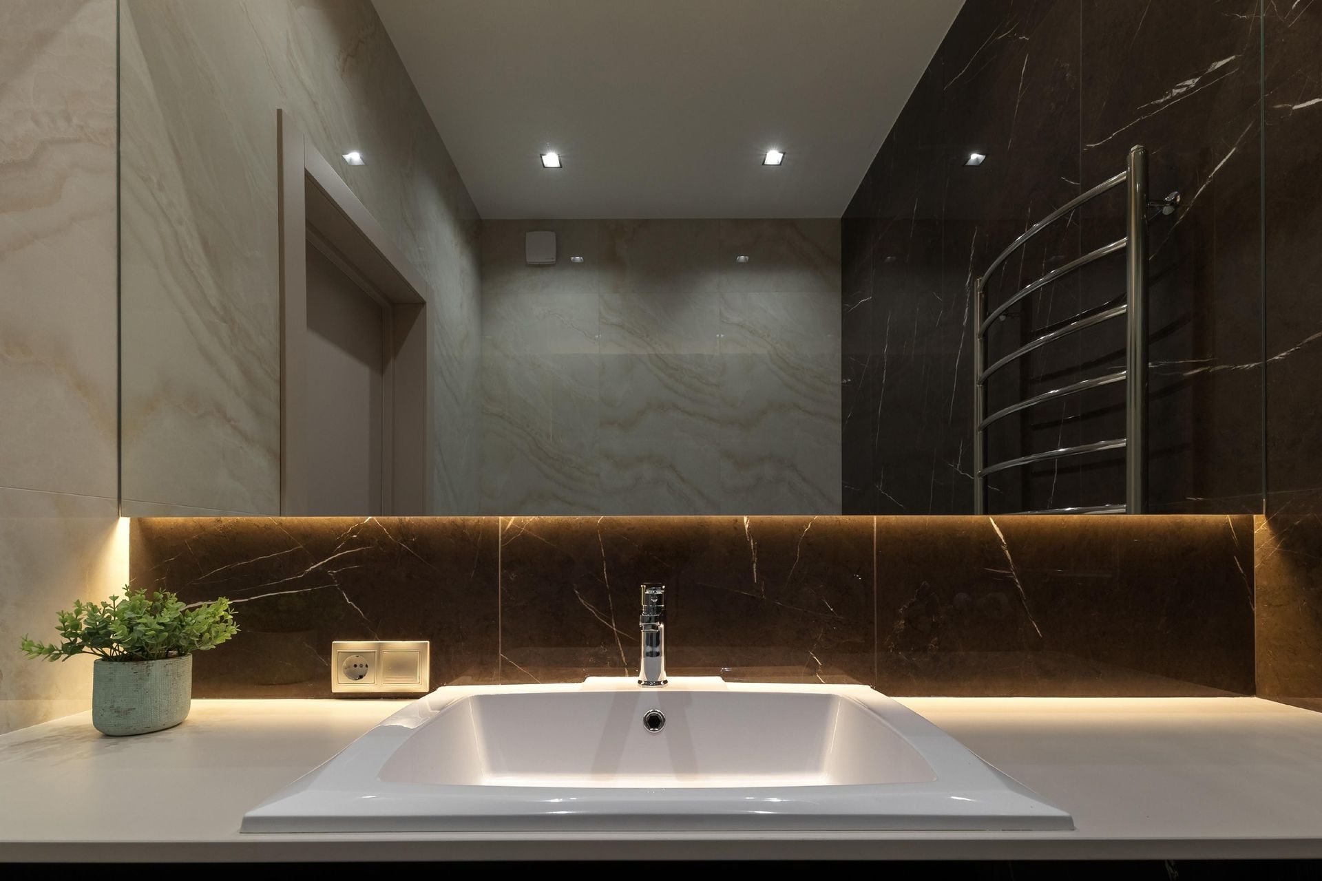 Modern bathroom vanity with white sink, backlit mirror, marble walls, and small plant from a front view