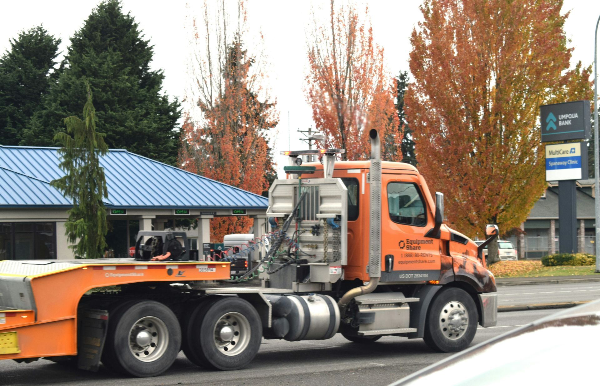 Orange truck with equipment on flatbed driving on a street near fall trees and a blue-roofed building.