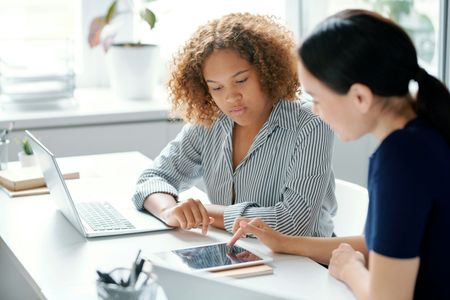 Two women working together at a desk; one points to a tablet screen while the other looks on.