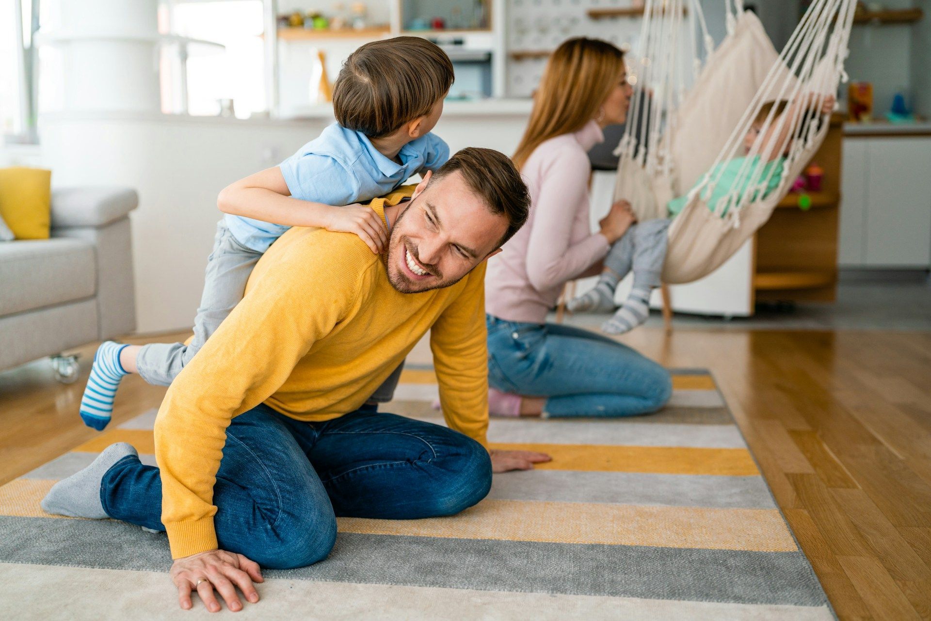Dad playing with son on rug, mom with baby in hammock, home setting.
