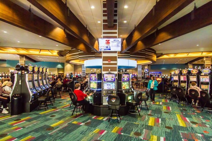 Wide casino floor with slot machines, colorful carpet, and a few seated players under bright ceiling lights
