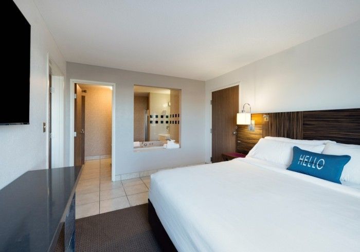 Modern hotel room with a king bed, blue accent pillow, bedside lamp, and bathroom doorway.