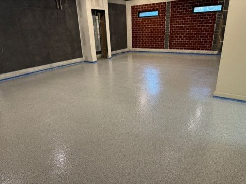 A hallway with polished dark brown concrete floors, light gray walls, and white trim. Unused cables are attached to the wall. Two windows are visible in the background.