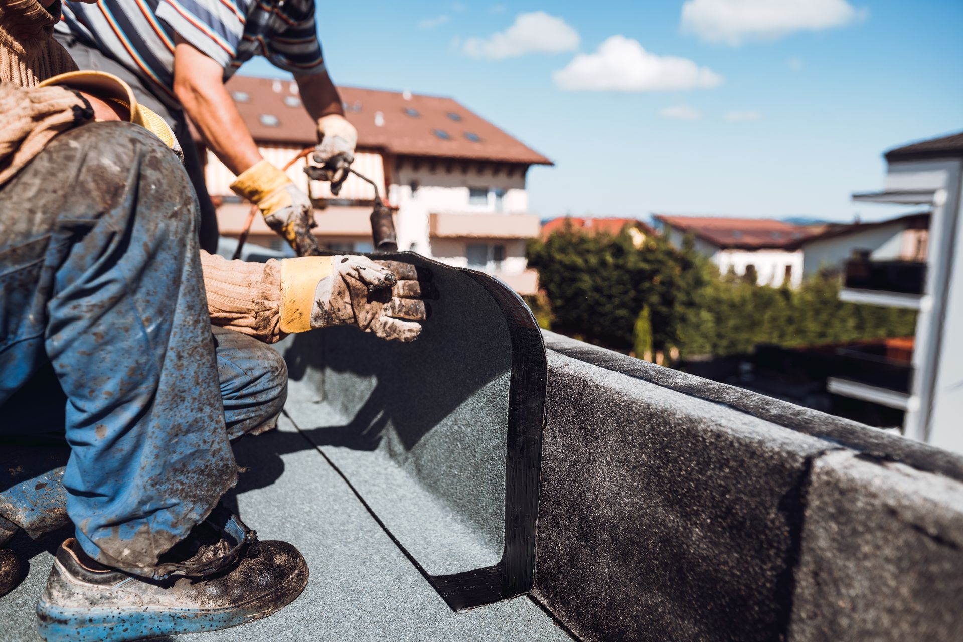 Workers installing roofing on a house, smoothing dark shingles along the roof edge.