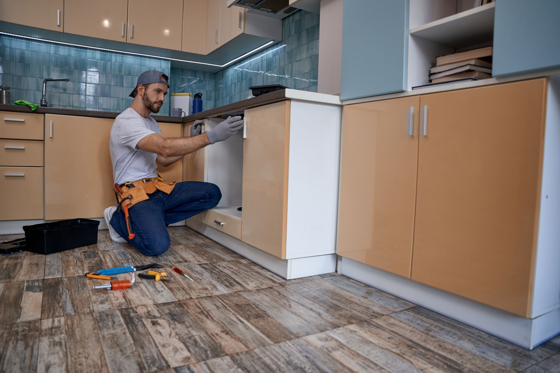 Person kneeling in a kitchen, installing white cabinets with tools on the floor.
