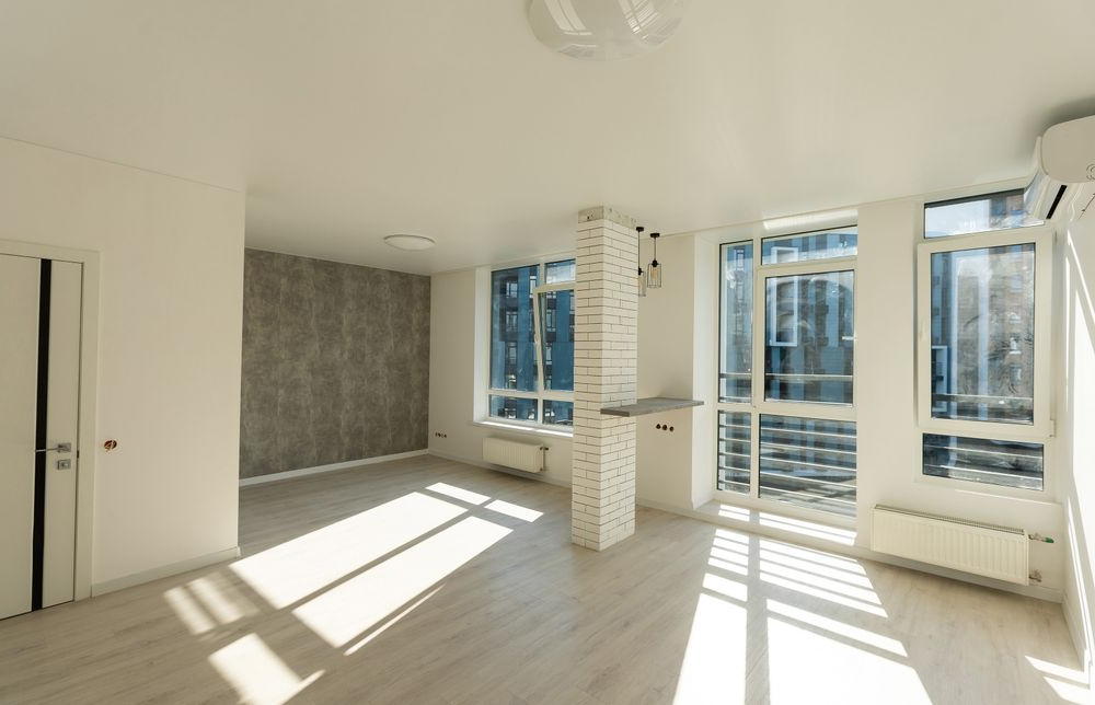 Bright empty room with large windows, white walls, and sunlight casting shadows on light wood floors