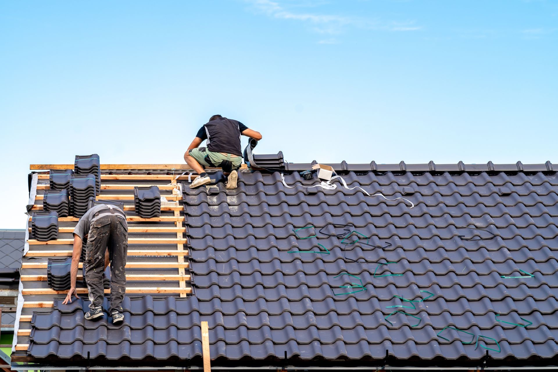 Roofers installing dark tiles on a house roof under a blue sky