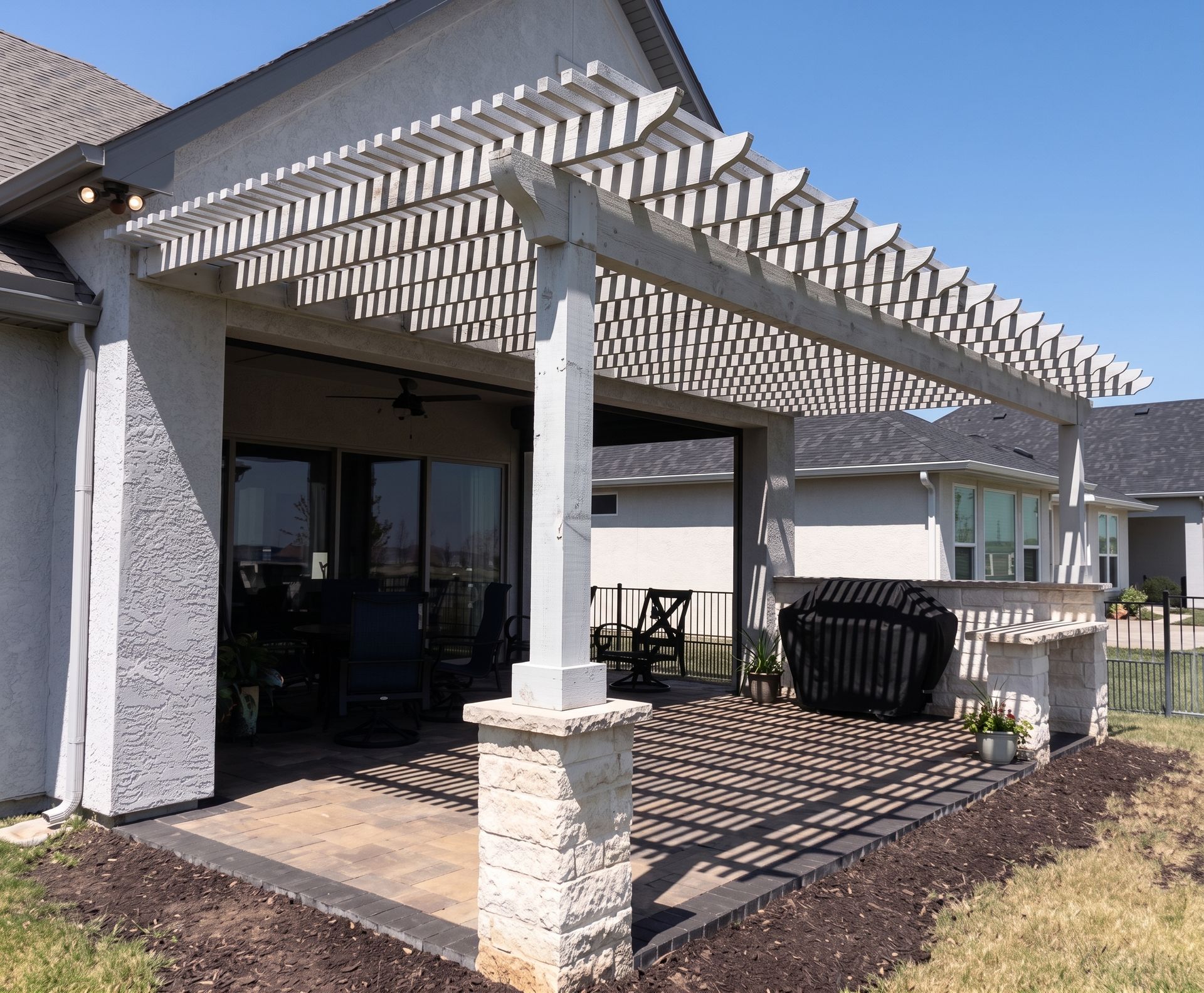 Patio with white pergola and stone columns beside a house, casting striped shadows on the deck.